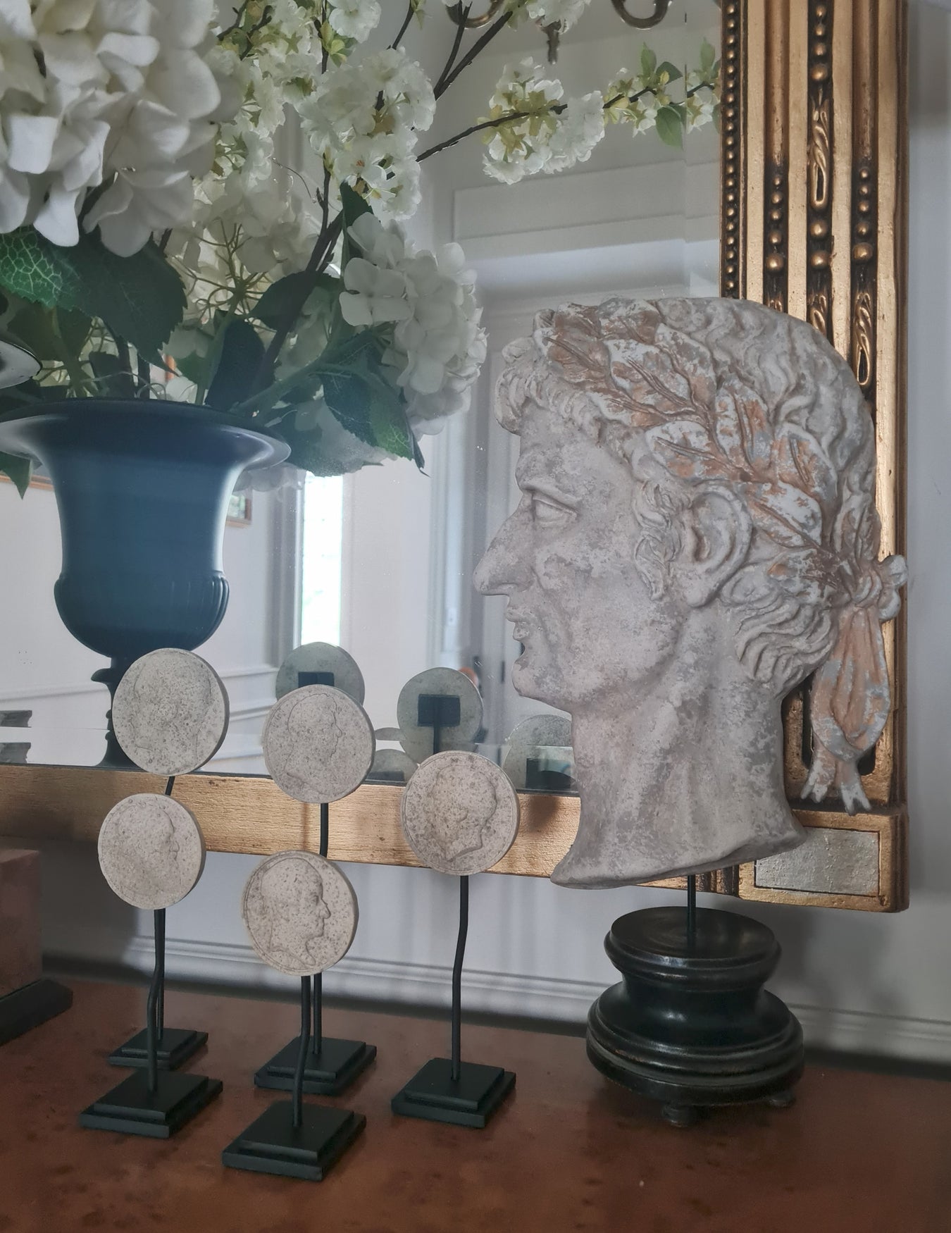 Decorative head sculpture and circular art pieces on a wooden surface with a mirror and flowers in the background.